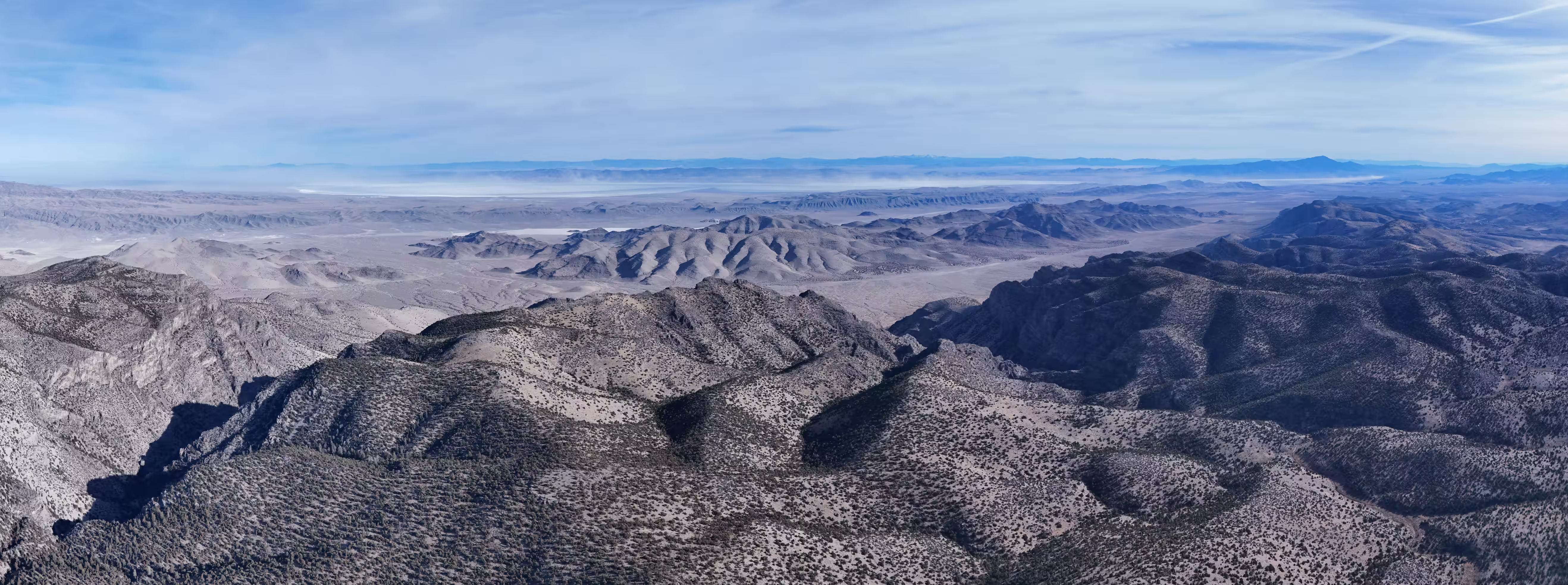 Panoramic aerial photograph of a vast desert mountain landscape under a pale blue sky with wispy clouds. In the foreground, rugged mountain ranges with sparse scrubby vegetation create a mottled gray-brown texture across steep ridges and shadowed valleys. The terrain extends into the middle distance where additional mountain ranges fade into atmospheric haze. On the far horizon, a large white dry lake bed (playa) is visible, bordered by distant mountain silhouettes. The image captures the characteristic Basin and Range topography of the American Southwest, emphasizing the immense scale and arid beauty of the region.​​​​​​​​​​​​​​​​