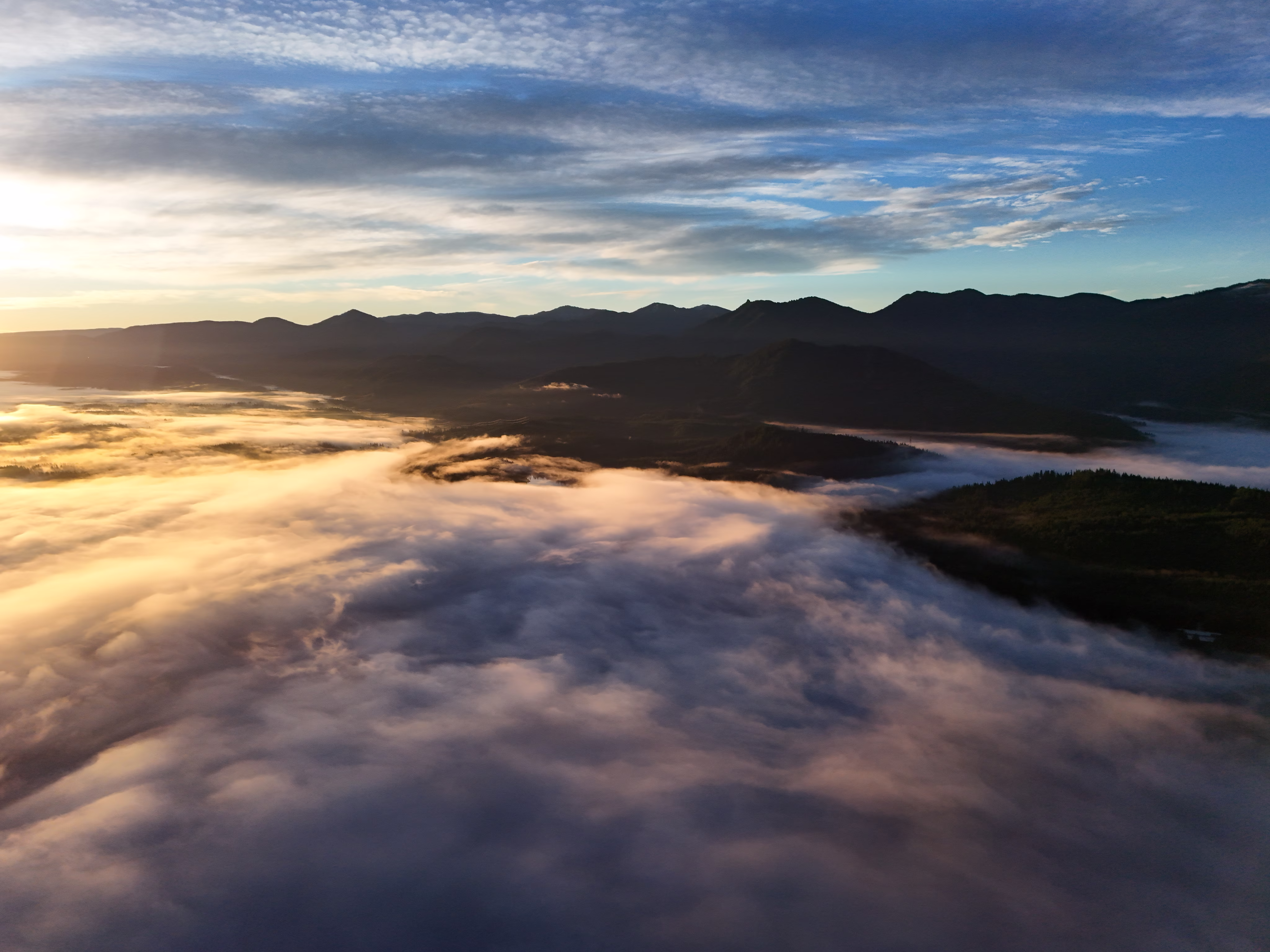 An aerial view above a sea of clouds filling a vast mountain valley in the early morning. A rugged mountain range with multiple sharp peaks creates a dramatic dark silhouette across the middle of the frame. Below, billowing white clouds with warm golden and peachy tones on the left side transition to cooler blue-white formations on the right, creating a three-dimensional effect. The clouds appear to flow and undulate around forested mountainsides. Above, a clear blue sky with wispy cloud streaks stretches across the top. The directional golden light suggests early morning or evening, highlighting the ethereal quality of the cloud inversion landscape.