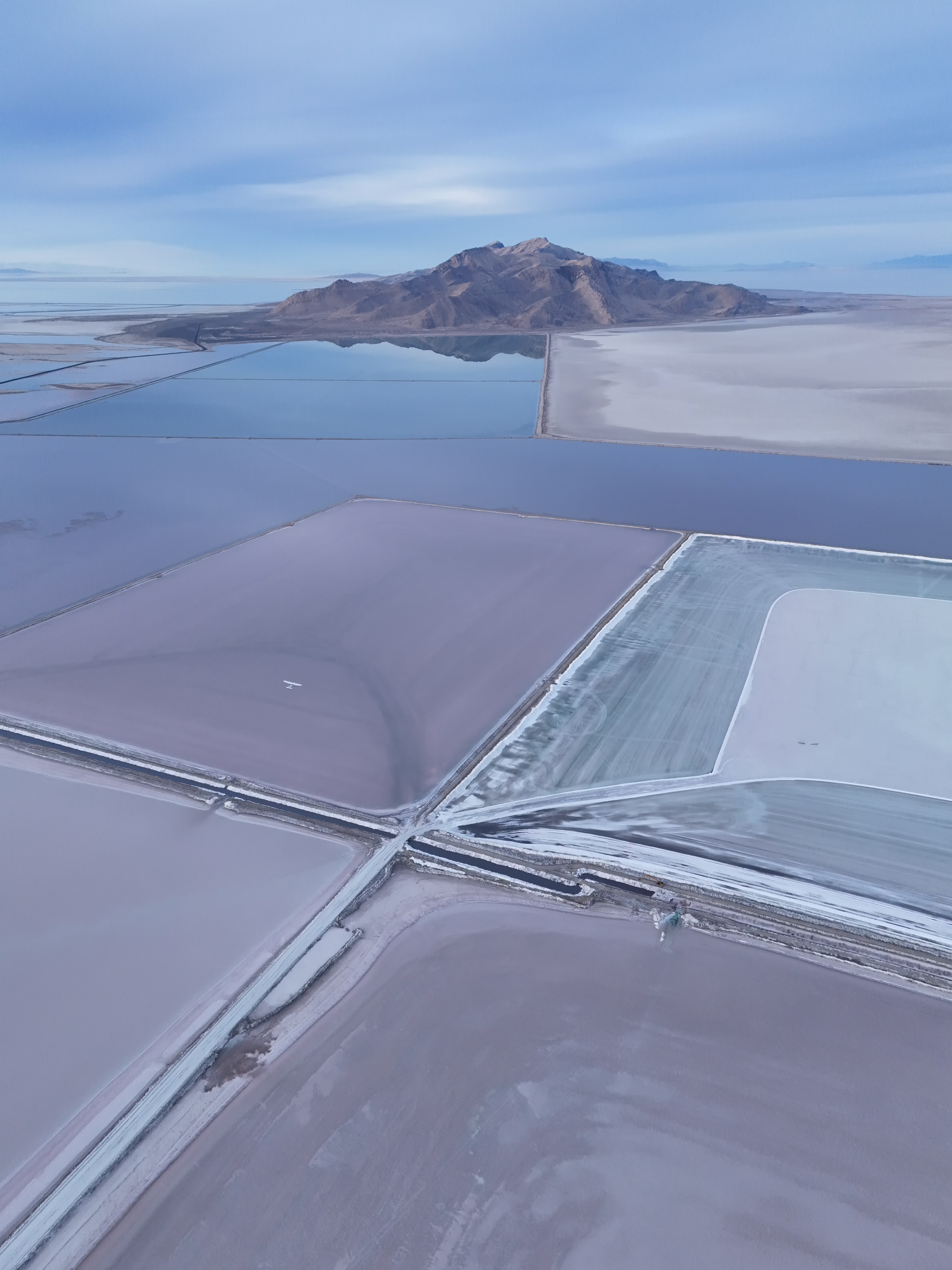 Aerial photograph looking down at salt evaporation ponds near the Great Salt Lake, Utah. A small white airplane is visible flying low over the ponds in the center of the frame, providing a striking sense of scale against the vast industrial landscape. The foreground features large rectangular ponds in muted lavender, mauve, and grayish-white tones, separated by white salt-crusted levees that meet at sharp angles. One pond on the right shows crystallized salt deposits forming delicate patterns along its edges. Beyond the ponds, a deep blue reservoir stretches toward a rugged brown mountain that rises dramatically from the flat lakebed, its peaks catching soft light. The background fades into pale salt flats and a hazy sky with thin, wispy clouds. The vertical composition emphasizes the geometric abstraction of the human-made ponds against the natural landforms.