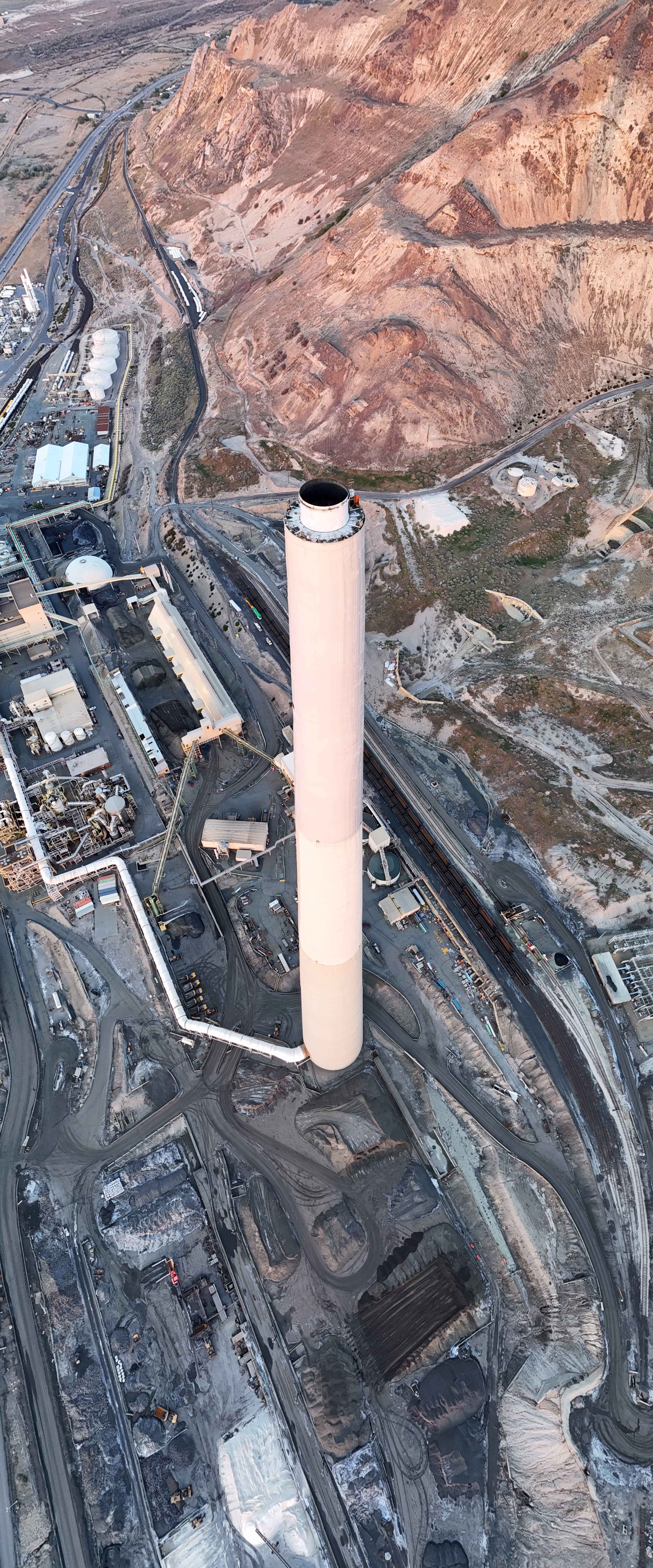 This high-angle aerial photograph captures a massive, solitary smokestack rising vertically from a sprawling industrial complex nestled at the base of arid, reddish-brown mountains. The smokestack, painted a pale cream or off-white, dominates the center of the frame, tapering slightly towards the top where a dark, circular opening is visible. At its base, a thick white pipe connects to the structure, snaking away towards a cluster of industrial buildings, storage tanks, and processing equipment to the left. The surrounding facility is a maze of gray infrastructure, including long conveyor belts, rail tracks, and piles of dark material scattered across the dusty ground. To the upper left, white spherical storage tanks and rectangular buildings are visible near a highway that runs parallel to the mountain range. The evening light casts long, soft shadows across the terrain, illuminating the rugged texture of the mountain slopes in warm, golden hues.