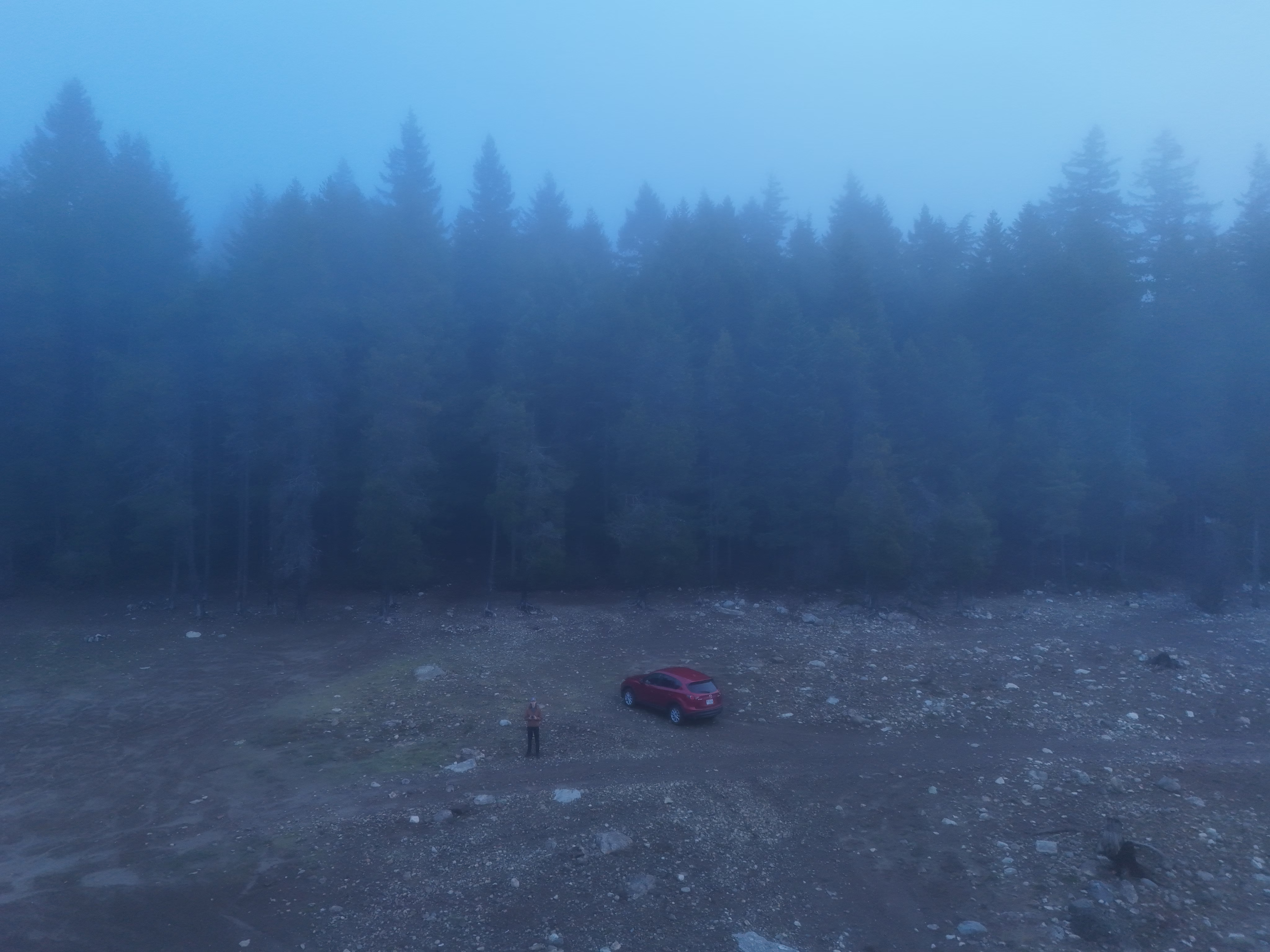 A drone's aerial view of a small clearing in a dense, foggy forest. A person stands next to a red car on a rocky, gravel-covered ground, both appearing small from the height of the drone. Tall coniferous trees form a thick wall at the back of the clearing, their silhouettes barely visible through heavy blue-gray mist and fog. The atmospheric conditions create a moody, minimalist scene with muted colors. Scattered rocks and patches of sparse vegetation cover the ground of the clearing. The composition emphasizes the isolation of the vehicle and person amid the vast, foggy wilderness.