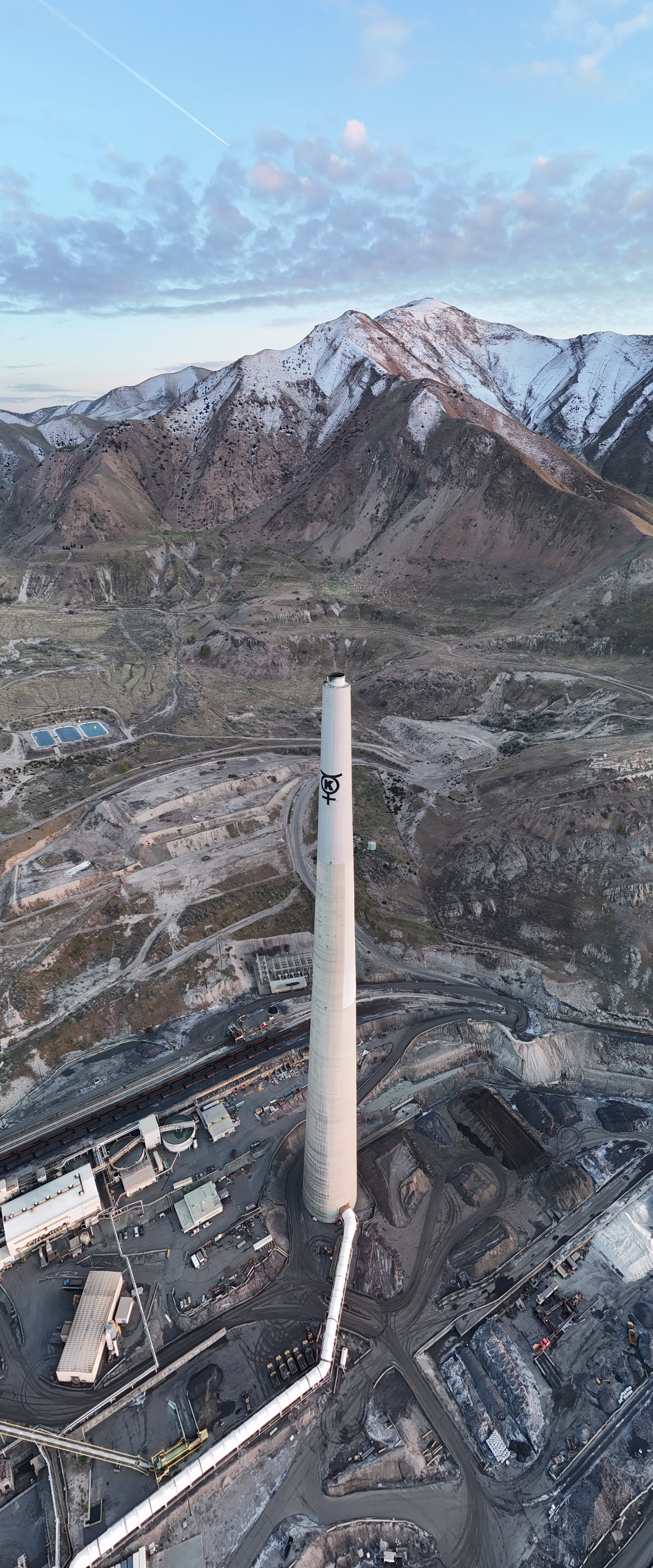 An aerial, high-angle vertical shot features a massive white concrete smokestack emblazoned with a black "K" logo near its summit, rising prominently from a sprawling industrial facility. At the base of the tower, a large white pipe curves along the ground, connecting to a network of low-lying white buildings, conveyor belts, and piles of dark grey material scattered across the dusty site. To the left of the main complex, three rectangular blue water retention ponds sit amidst the arid, brownish terrain. The background is dominated by a rugged mountain range with steep, rocky slopes partially covered in snow, particularly near the peaks which catch the warm, soft light of a low sun. Above, the sky is a pale blue filled with scattered, fluffy clouds and a thin white contrail cutting diagonally across the upper left. The lighting suggests late afternoon or early evening, casting soft shadows across the industrial grounds while illuminating the mountain ridges with a golden hue.